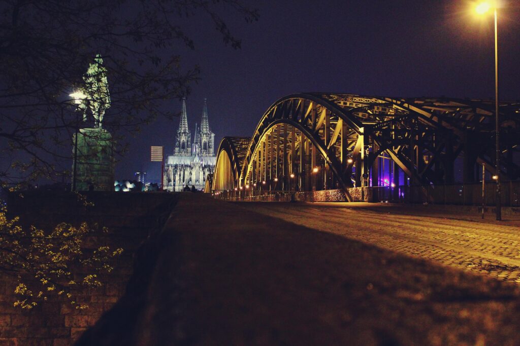 Illuminated view of Cologne Cathedral and Hohenzollern Bridge under evening lights in Cologne, Germany.